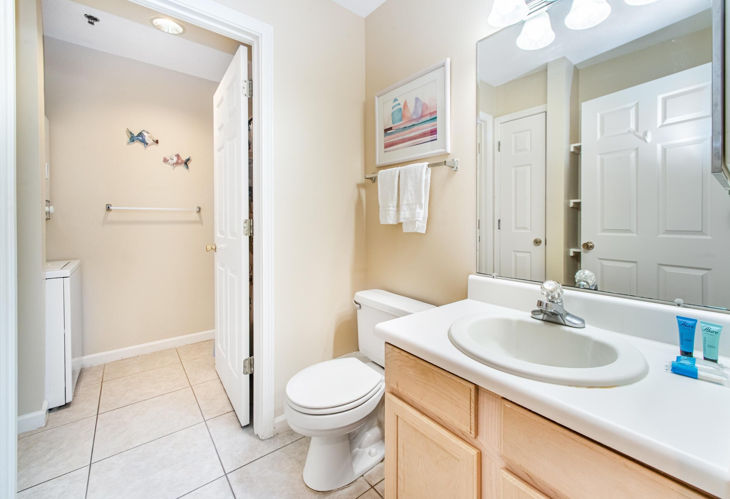 A bathroom with a toilet, sink, and mirror. There is a towel rack above the sink and a small framed picture on the wall.