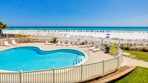 Une piscine entourée d’une clôture blanche, bordée de chaises de plage et de parasols, avec une plage et l’océan en arrière-plan.