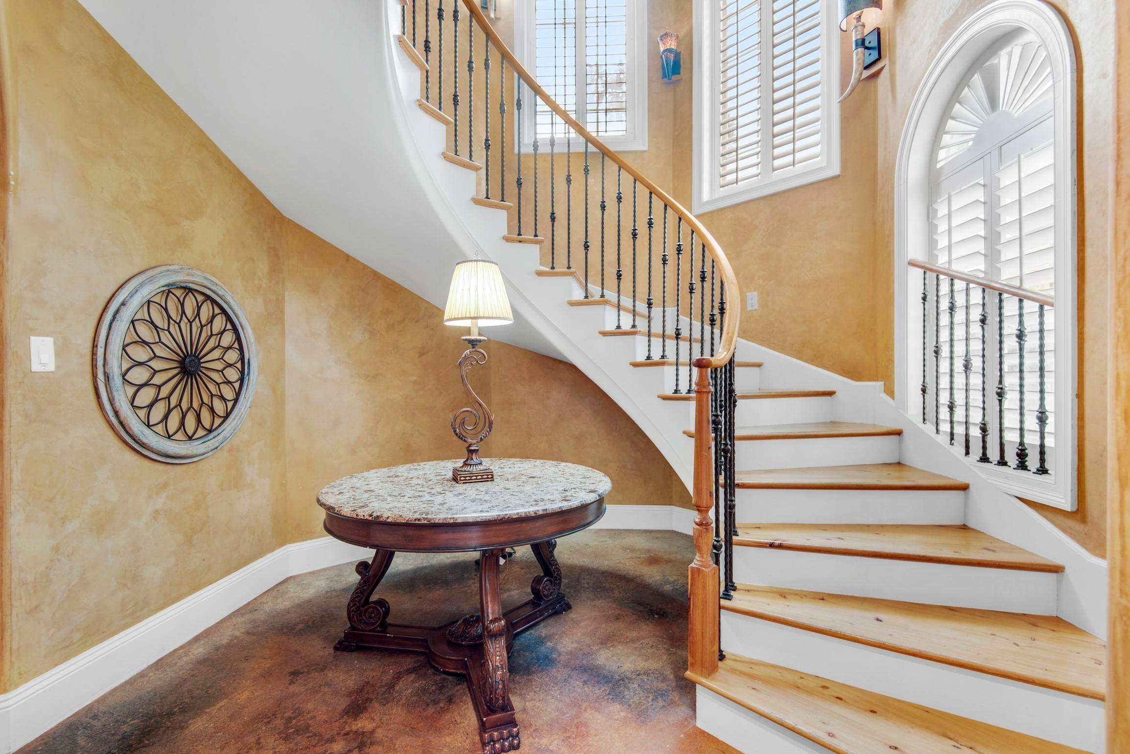 A grand staircase with wooden steps and black balusters, leading to a landing with a round table and a wall-mounted lamp.
