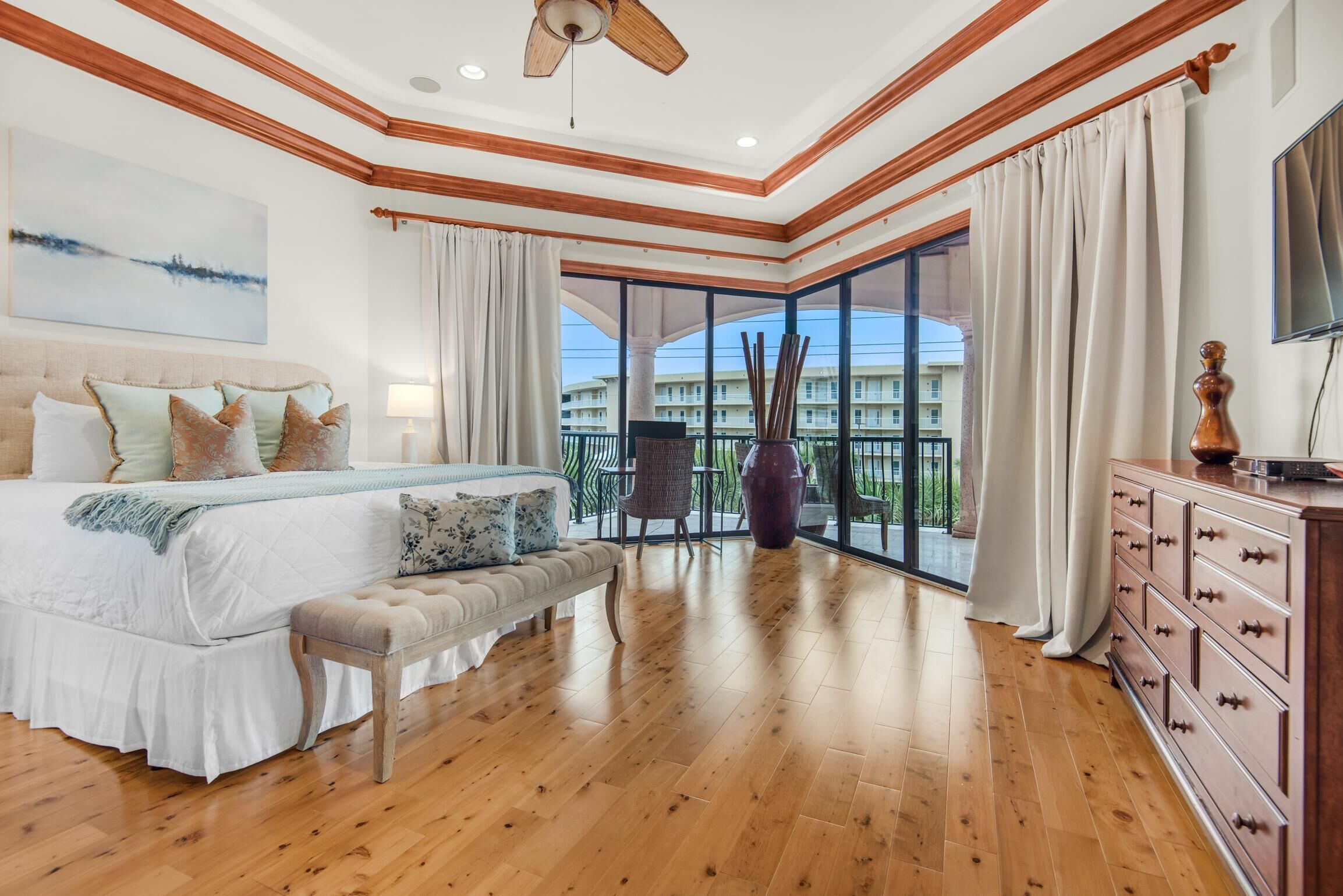 A bedroom with a large bed, wooden flooring, a ceiling fan, and a view of a building through large windows.