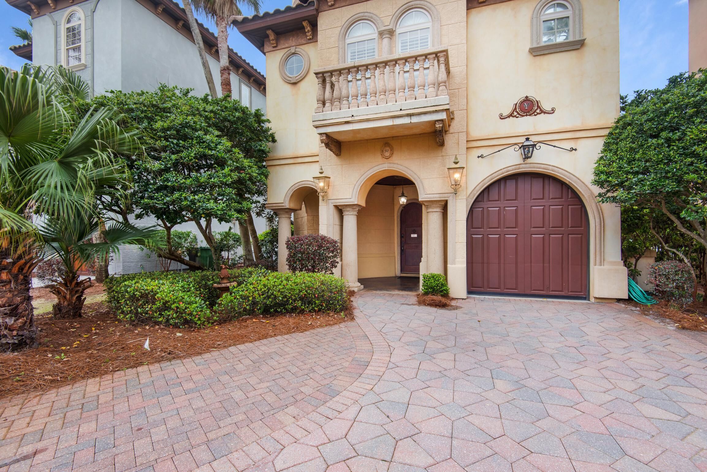 A two-story building with a large brown garage door, a balcony with ornate railings, and a well-maintained garden with palm trees and shrubs.