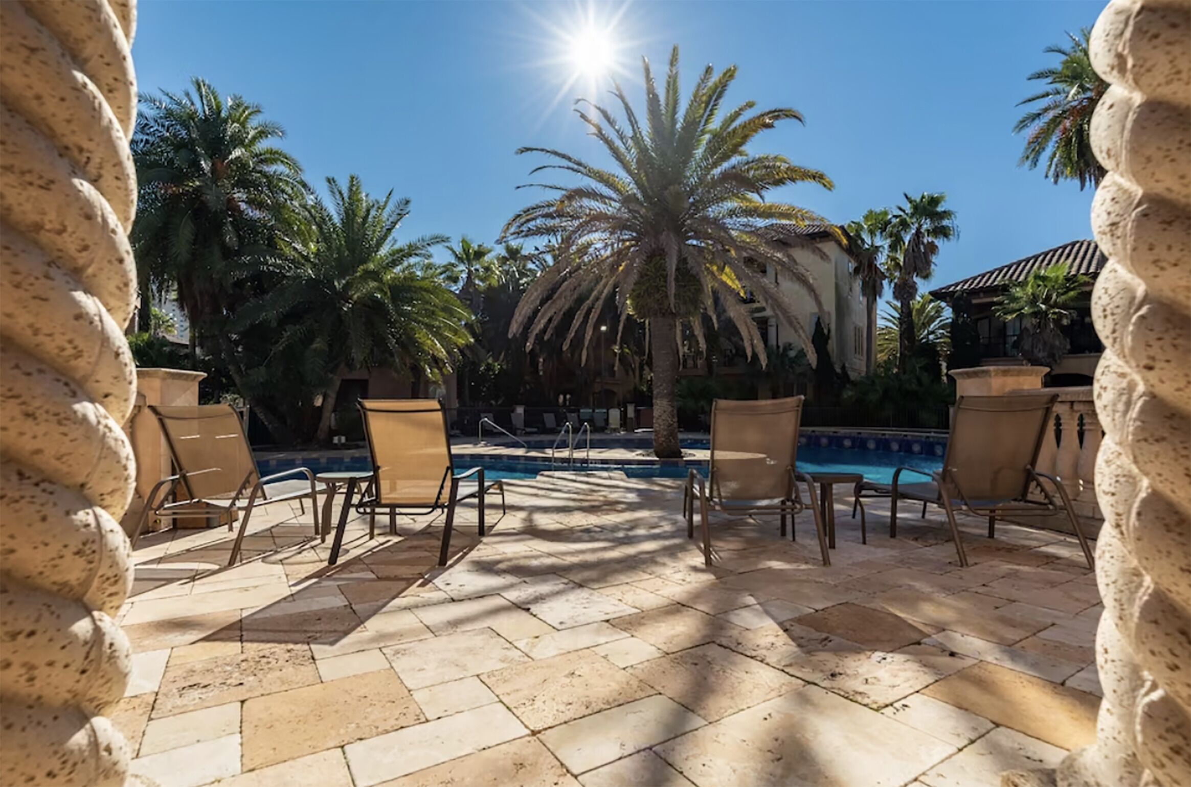 A poolside area with chairs and palm trees, a clear sky, and a building in the background.