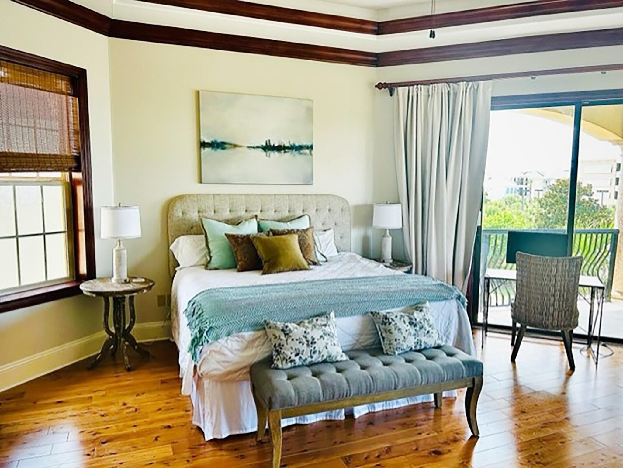 A bedroom with a large bed, two bedside tables, a bench, a view of the outdoors, and a painting on the wall.