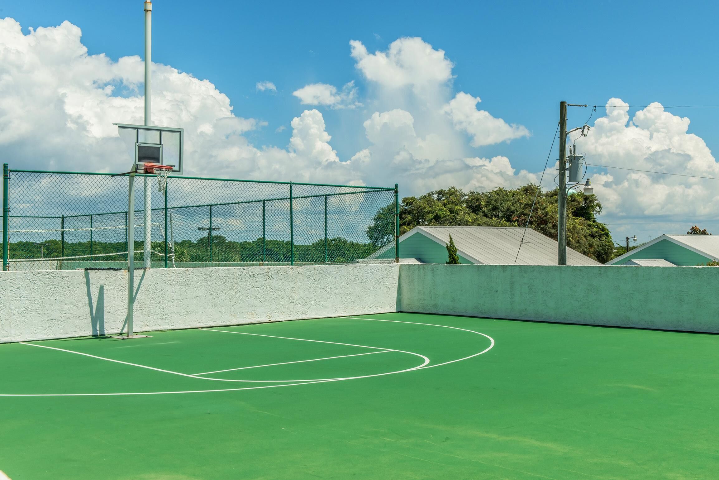Ein Tennisplatz im Freien mit einem Basketballkorb, umgeben von einem grünen Zaun und einer weißen Mauer.