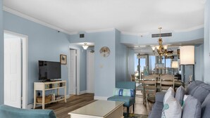 A living room with a television, a coffee table, and a sofa. There is a dining area with a table and chairs, and a kitchen in the background. - Surfside Resort #309 (Miramar Beach)