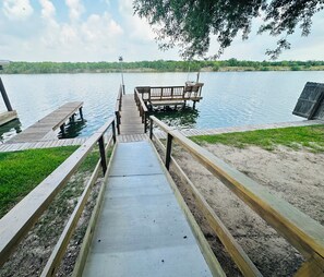 A wooden dock leading to a bench by a lake with a grassy area on one side and a sandy area on the other.