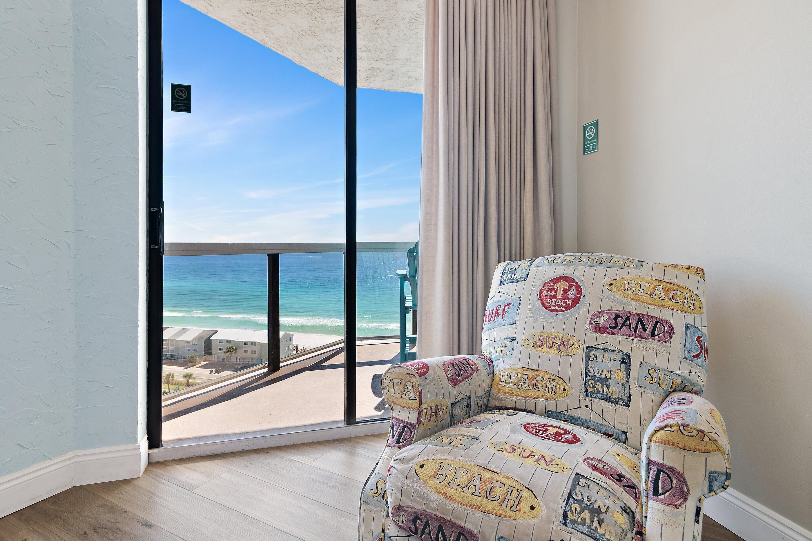 A room with a large window overlooking a beach, a patterned armchair, and a view of the ocean.