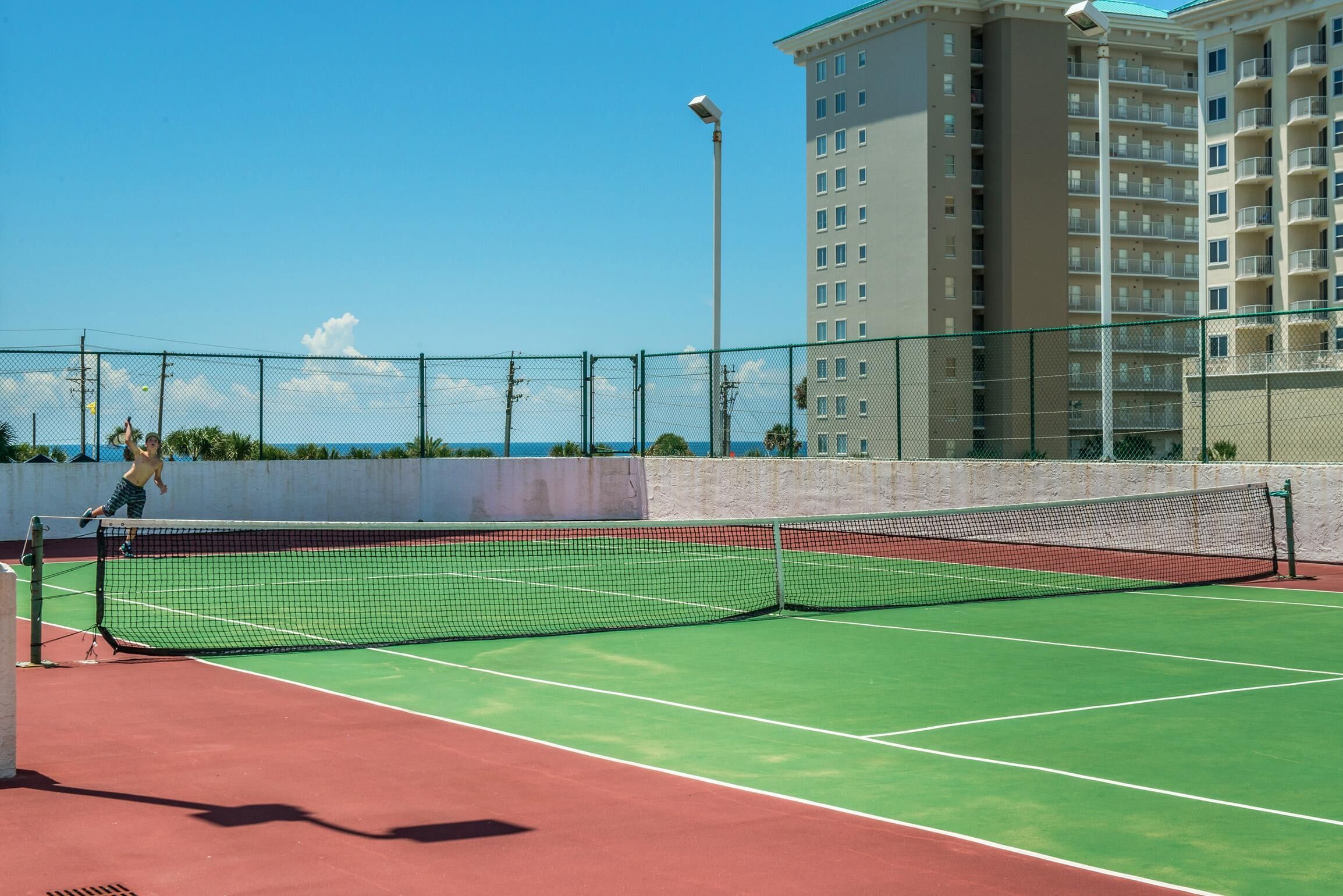 A tennis court with a net, surrounded by a fence, and a building in the background.