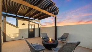 Eine Terrasse im Freien mit Feuerstelle und Korbstühlen, einem Pergola und Blick auf den Sonnenuntergang.