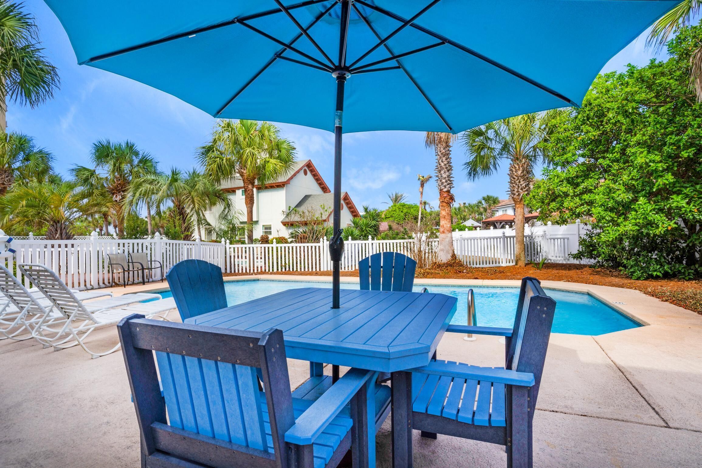 Een zwembadterras met een blauwe tafel en stoelen, een grote parasol en een witte hek in de achtergrond.