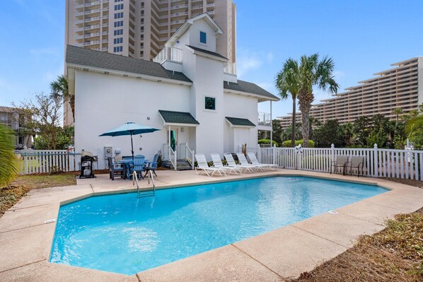 A swimming pool with lounge chairs and a table, surrounded by a white fence and palm trees.