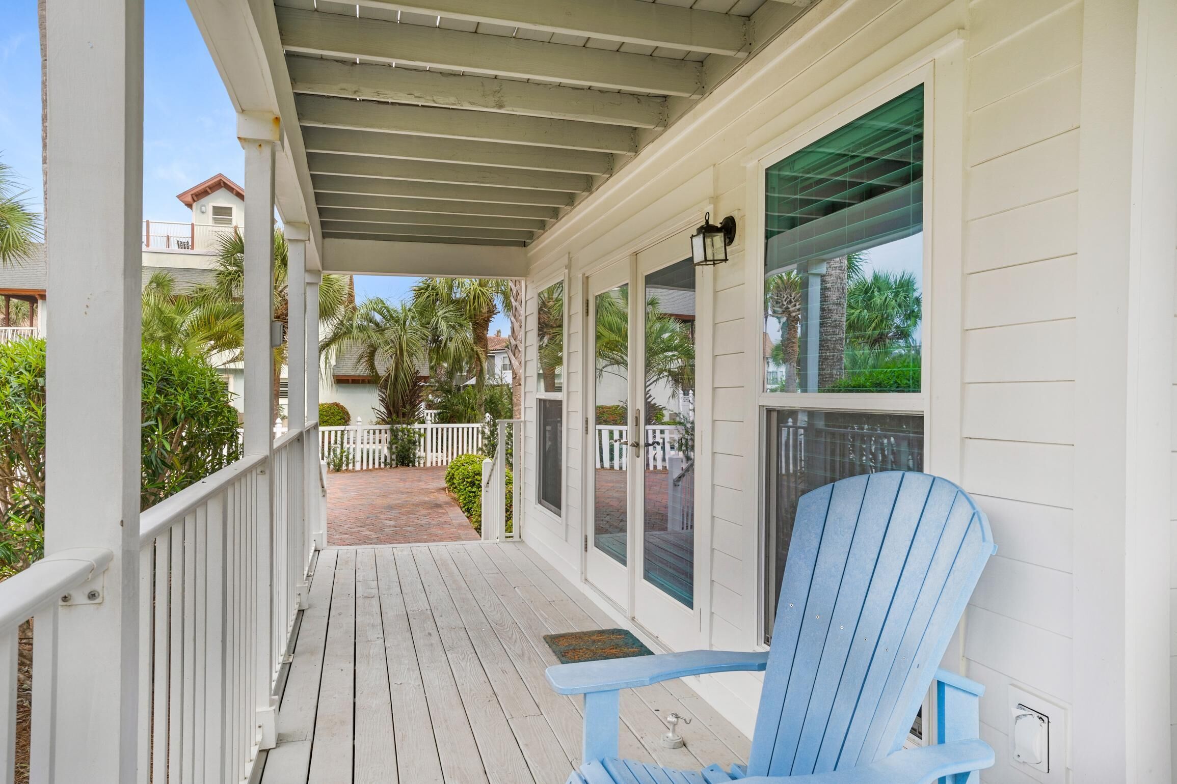 A covered porch with a blue Adirondack chair, white railings, and a view of a white house with a red roof in the background.