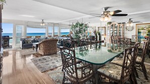 Un vaste espace de vie comprenant un coin repas avec une table à plateau en verre entourée de chaises en bois, un canapé et une vue sur l’océan.