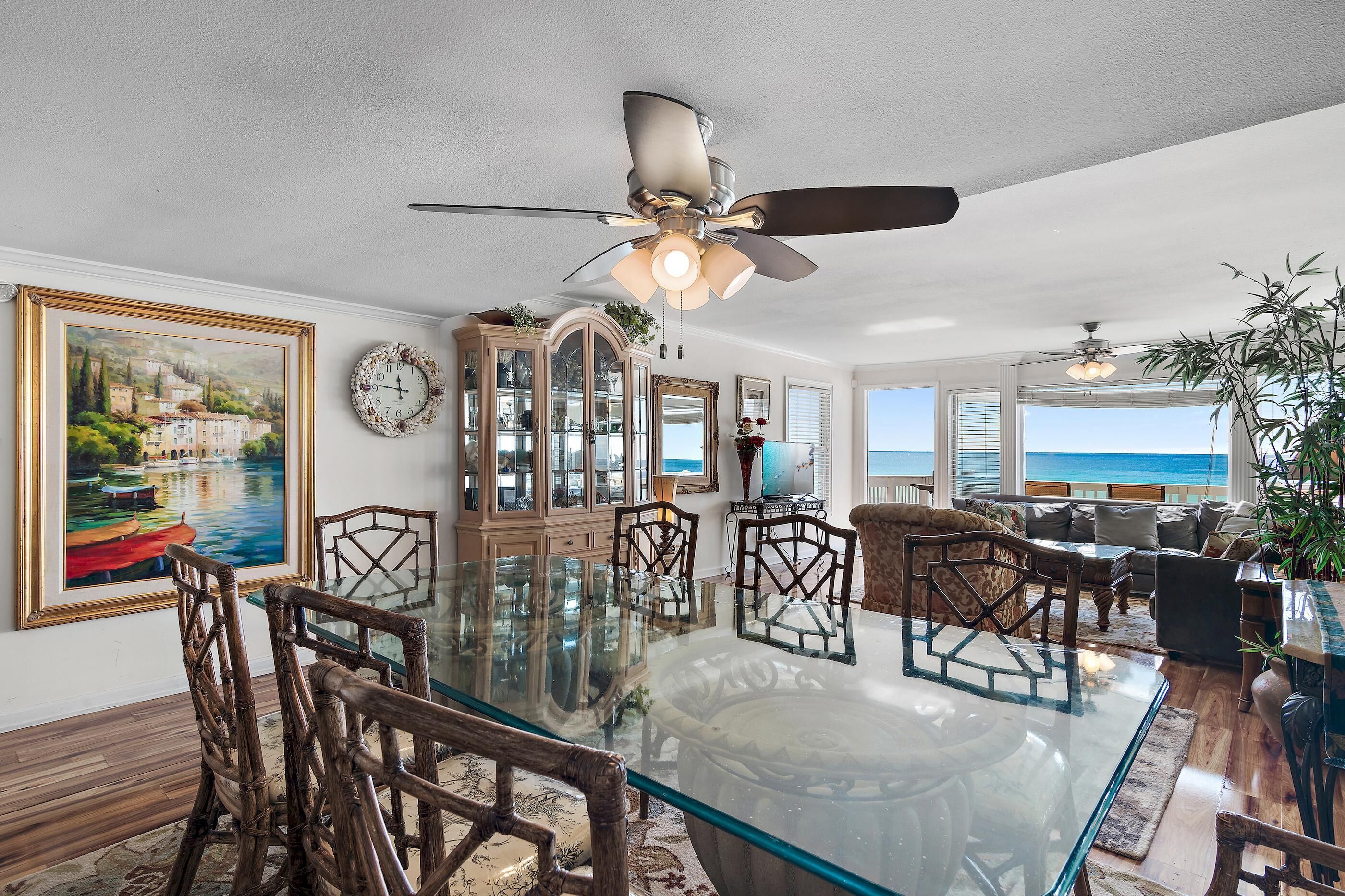 A dining area with a glass table and wooden chairs, a ceiling fan, and a view of the ocean.