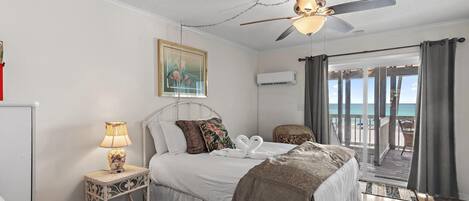 Un dormitorio con cama, ventilador de techo, mesita de noche con lámpara, un cuadro y vista a la playa desde un balcón.