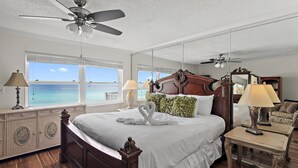 Une chambre avec un grand lit, deux lampes de chevet, un ventilateur de plafond et une vue sur l’océan grâce à de grandes fenêtres.