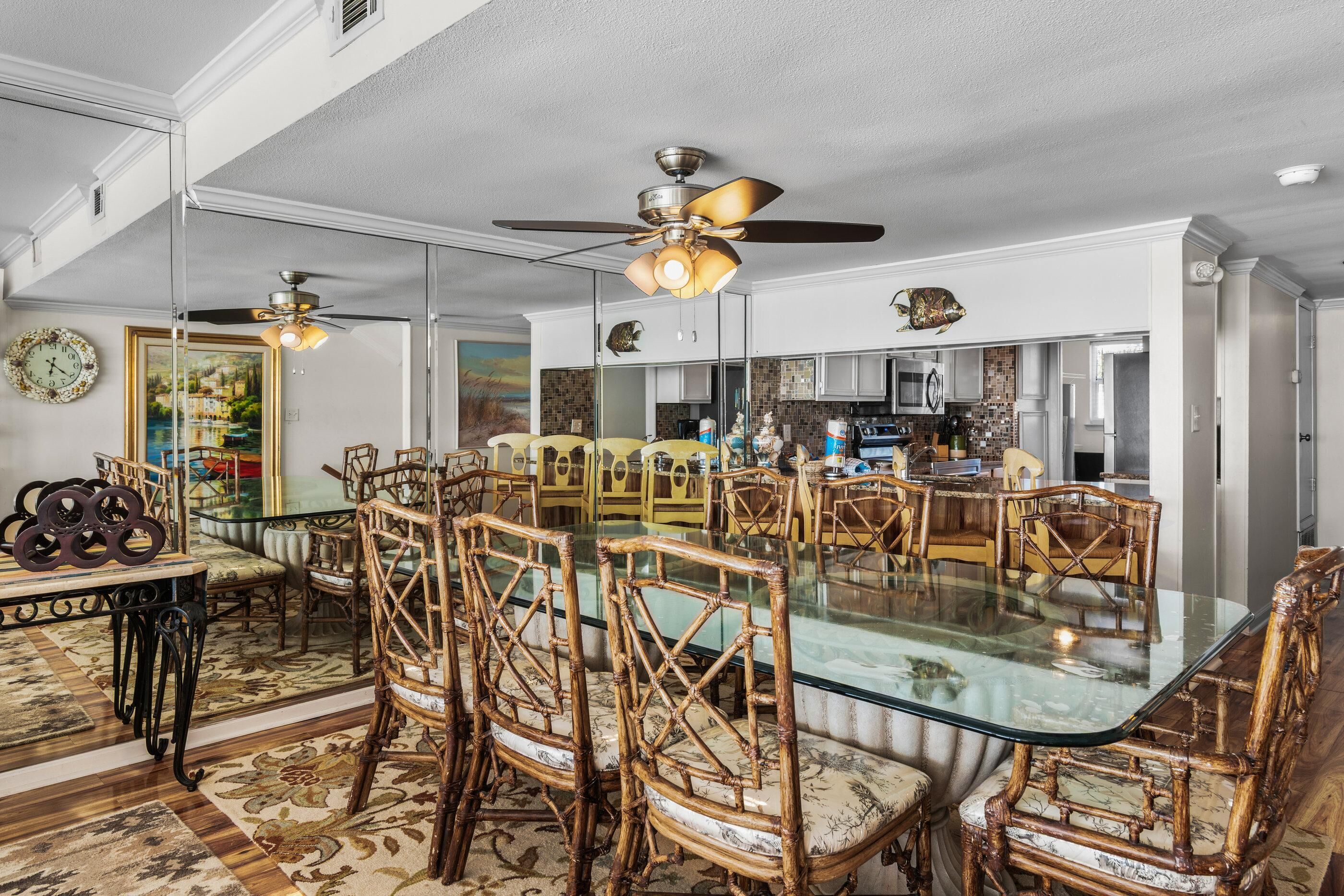 Un coin repas avec une table en verre entourée de chaises en osier, un ventilateur de plafond et une cuisine en arrière-plan.