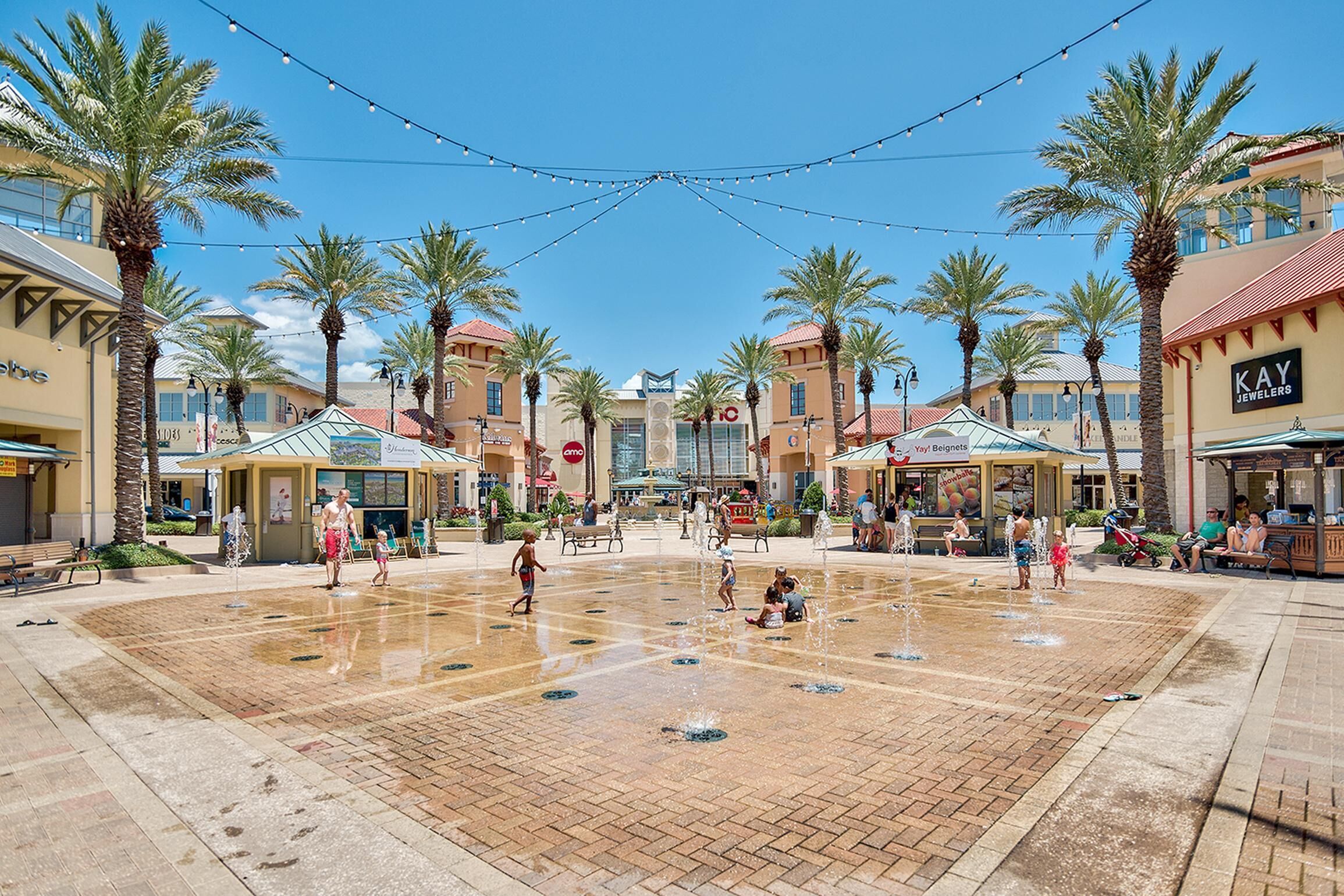 A public square with a water fountain, palm trees, and various shops.