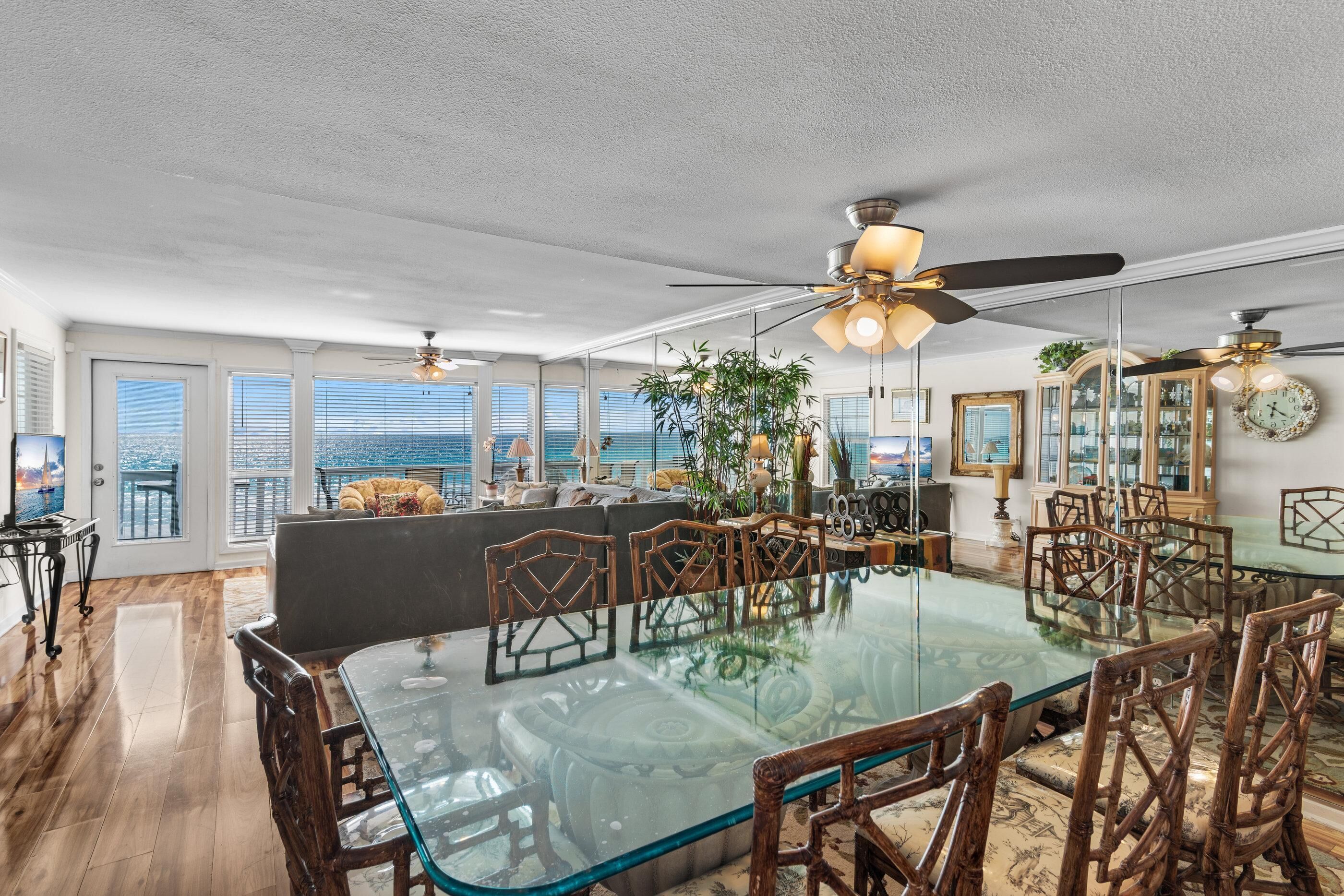 A spacious dining area with a glass-top table and wooden chairs, a ceiling fan, and a view of the city through large windows.