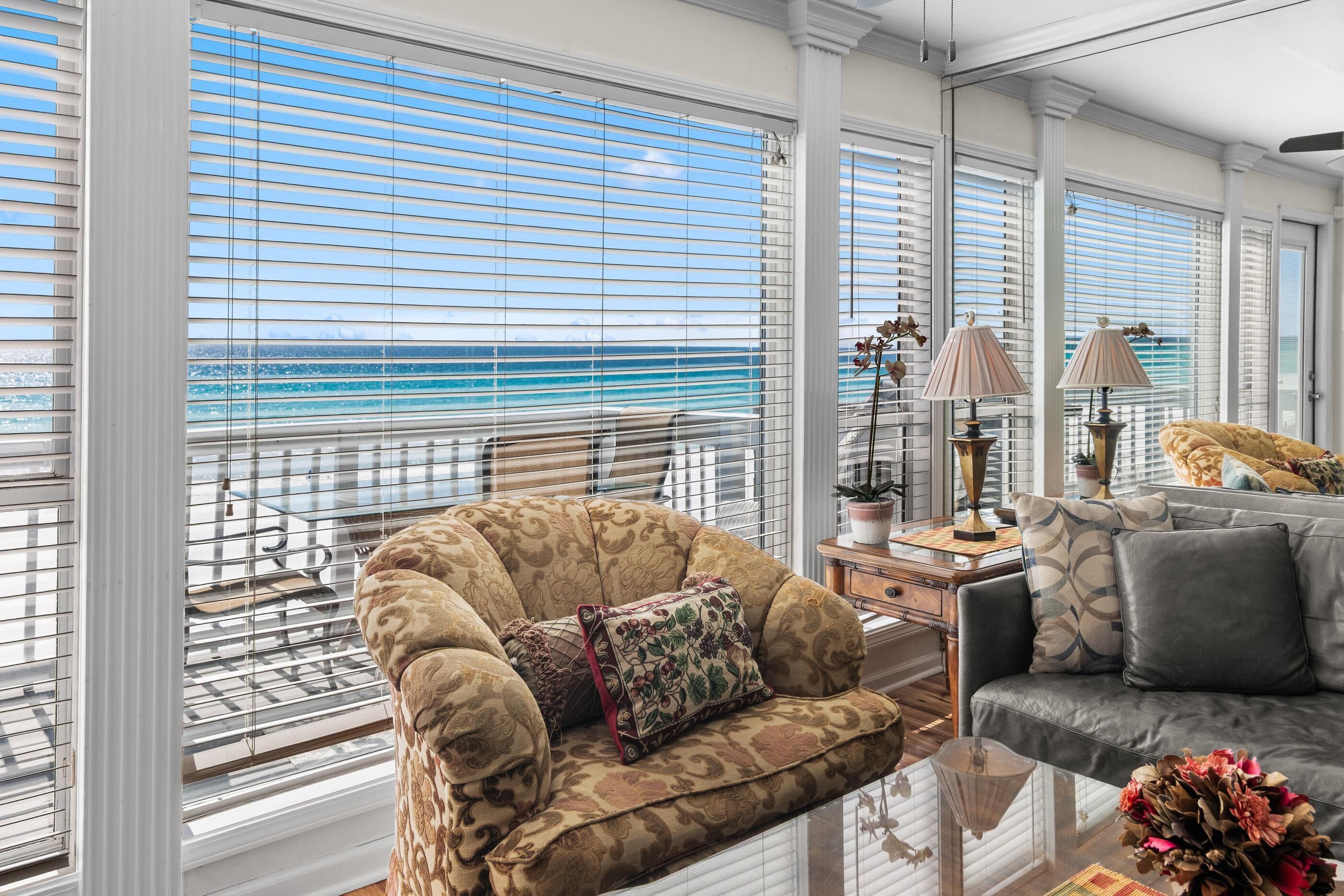 A spacious living room with a view of the ocean, featuring a patterned armchair, a sofa, and a side table with lamps and decorative items.
