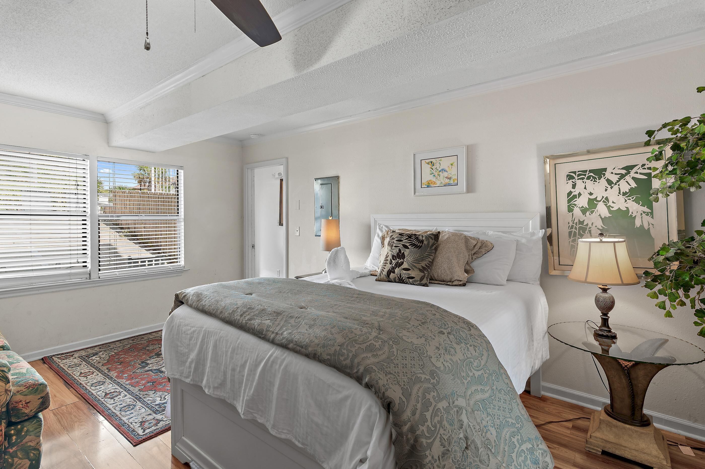 A bedroom with a large bed, a bedside table with a lamp, a window with blinds, and a door.