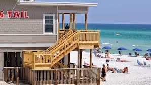 Un bâtiment situé en bord de mer, doté d’un escalier en bois menant à une terrasse, entourée de chaises de plage et de parasols.