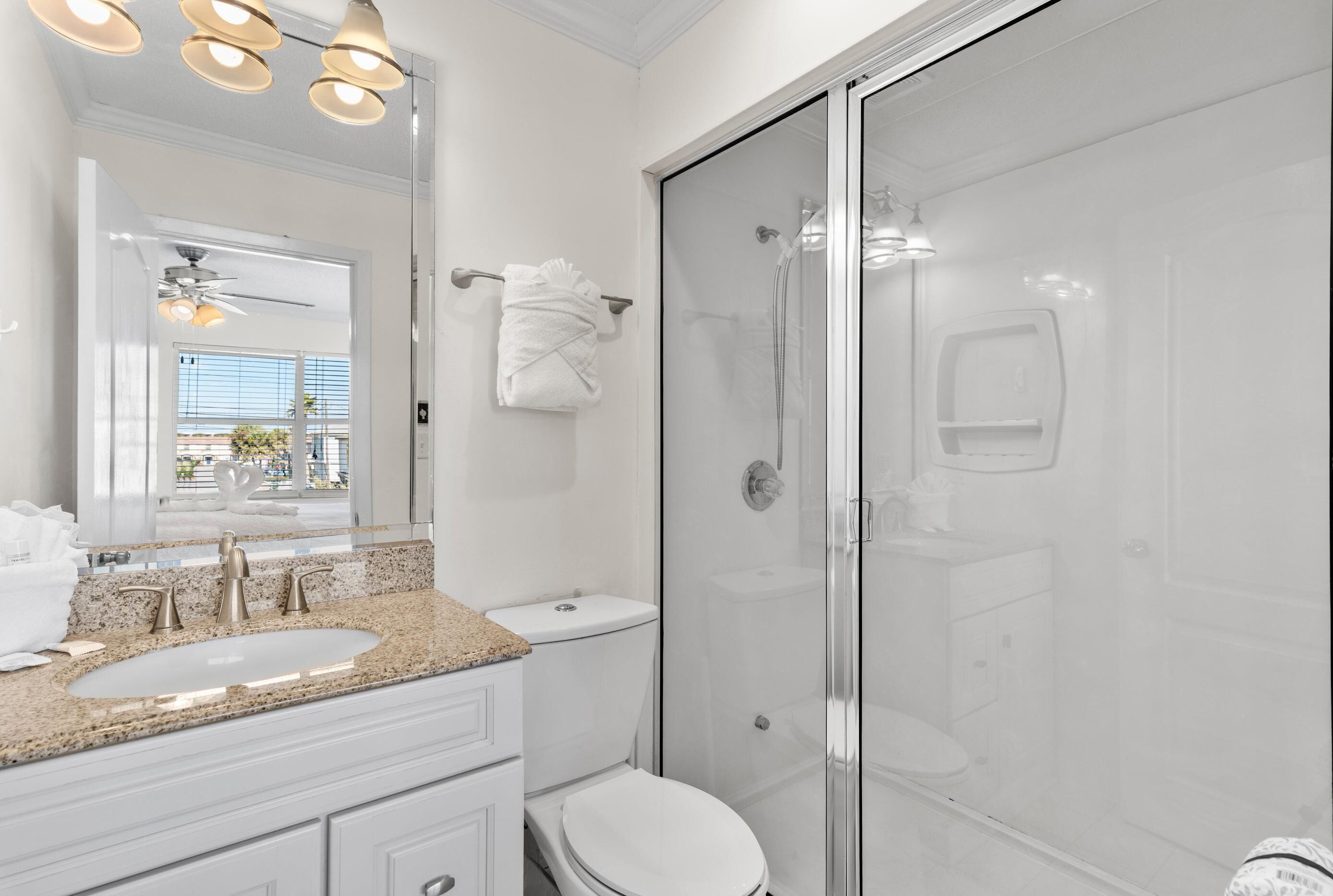 Une salle de bain moderne avec un lavabo surmonté d’un plan en granit, des toilettes blanches, une douche en verre et un grand miroir avec des luminaires au plafond.