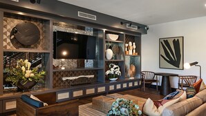 A modern living room with a large flat-screen TV, built-in shelving with decorative items, a comfortable sofa, and a dining area with a table and chairs.