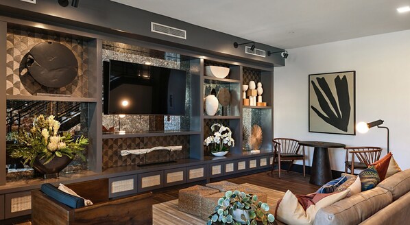 A modern living room with a large flat-screen TV, built-in shelving with decorative items, a comfortable sofa, and a dining area with a table and chairs.