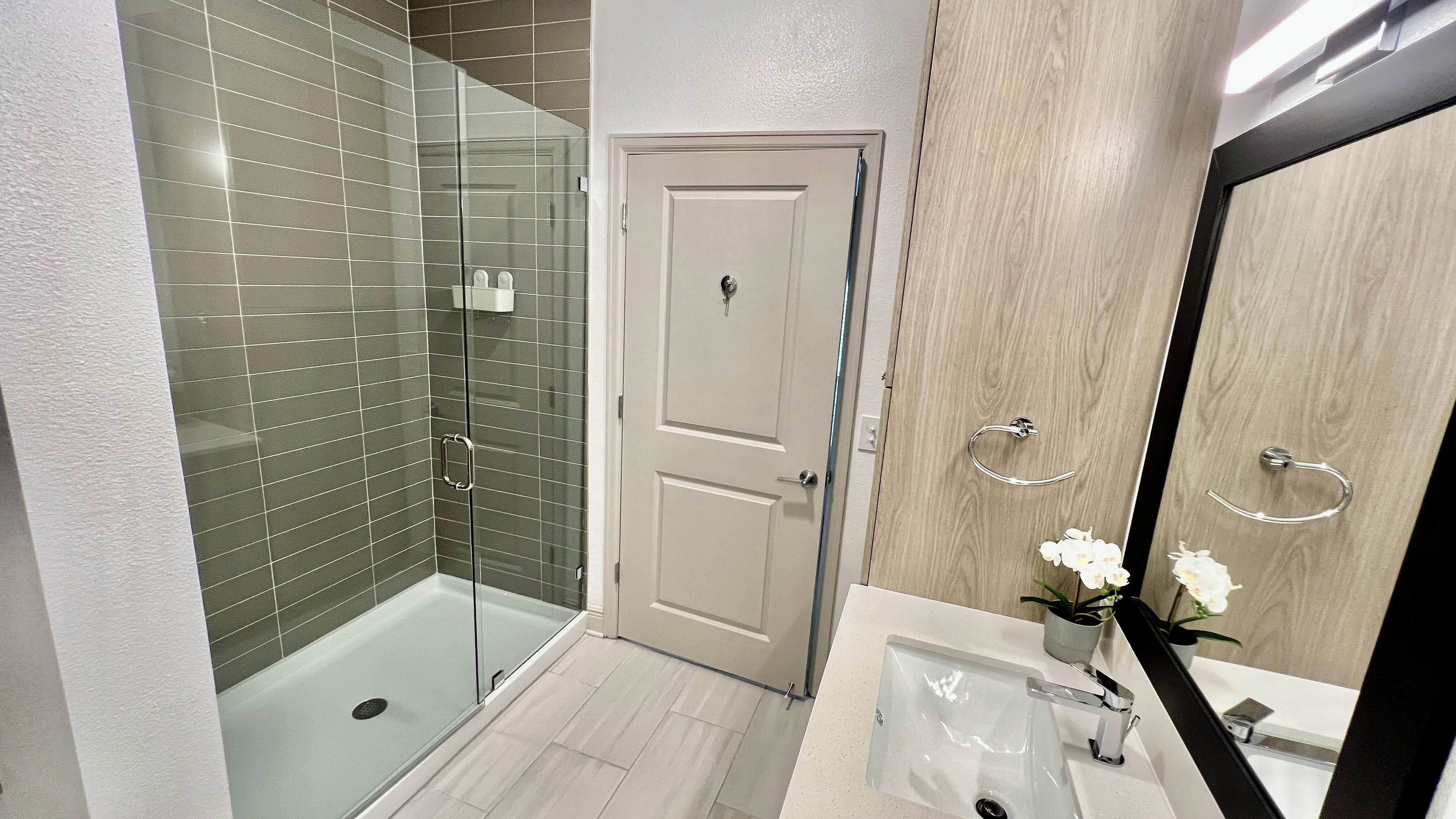 Une salle de bain moderne avec une douche à parois de verre, une porte blanche, un meuble-lavabo en bois et un miroir agrémenté d’un porte-serviette.