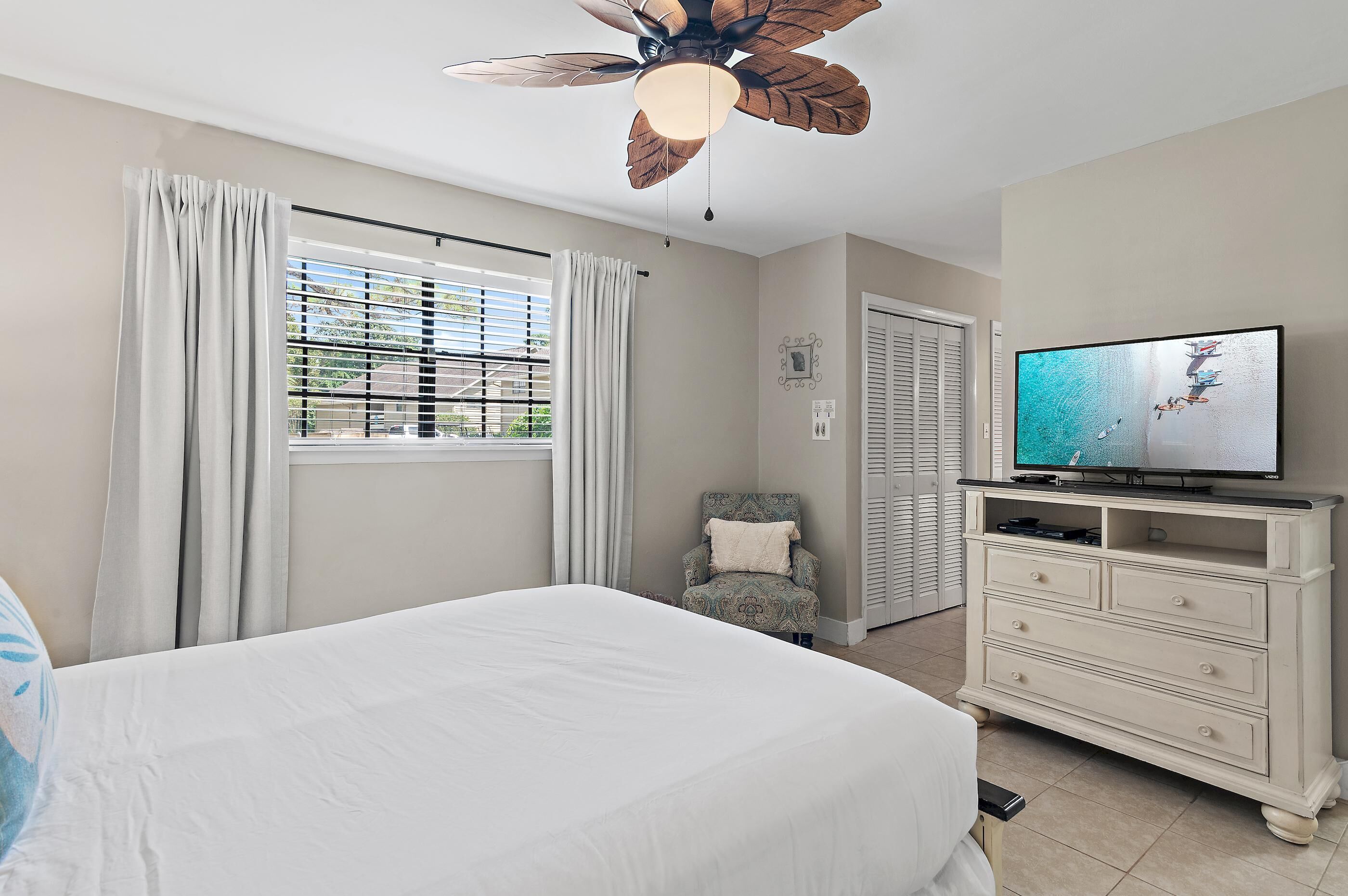 A bedroom with a bed, a television on a dresser, a ceiling fan, and a window with curtains.