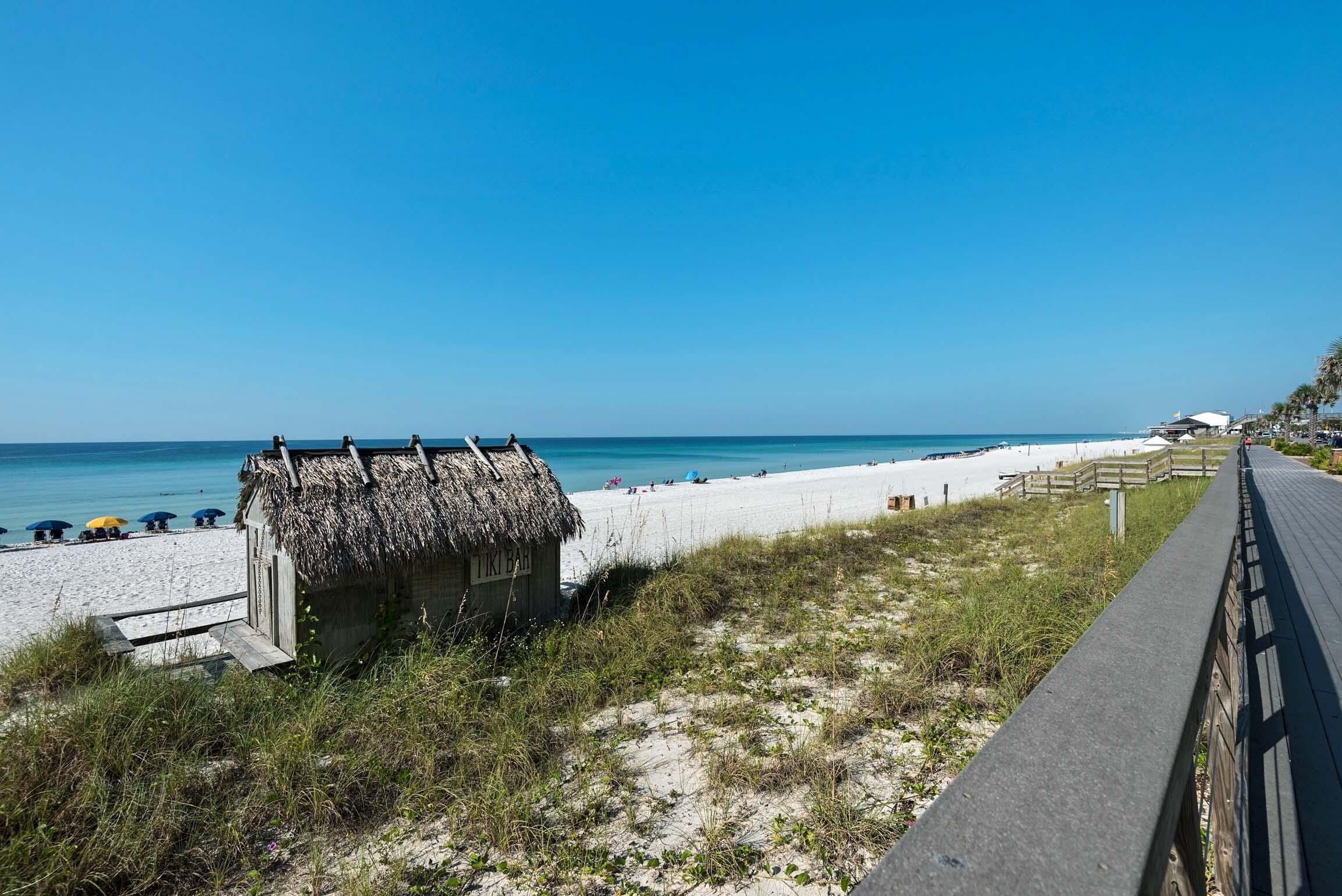 En strand med en liten stuga med halmtak, där det står ”TIKI BAR”, och en träpromenad som leder ner till stranden.