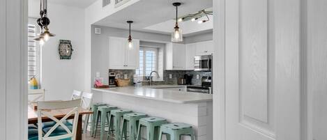 Una cocina moderna con armarios blancos, una isla grande con taburetes de bar, iluminación colgante y un reloj en la pared.
