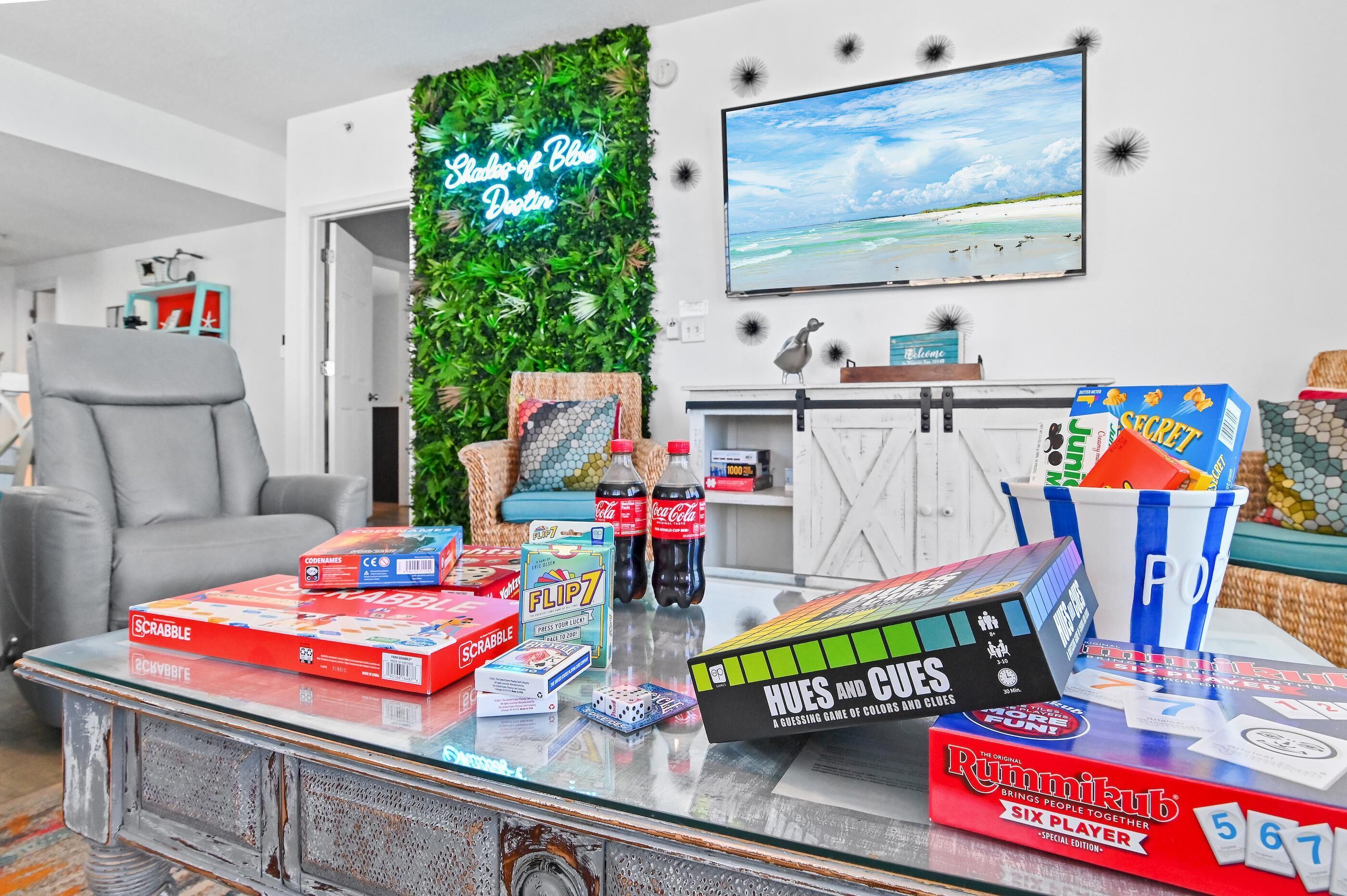 A living room with a glass coffee table holding various board games, a neon sign, and a TV displaying a beach scene.