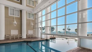An indoor swimming pool with clear views of the ocean through large windows, a tiled floor, and several chairs along the poolside. - Majestic Sun 1012a (Miramar Beach)