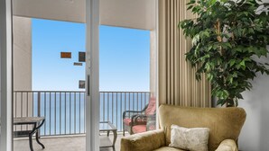 A room with a sliding glass door leading to a balcony with a view of the sea. There is a beige armchair with a pillow and a potted plant to the right. - Majestic Sun 1012a (Miramar Beach)