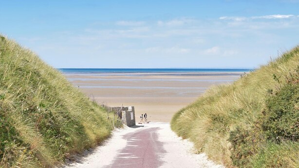 Ein Pfad, der zu einem Strand mit Dünen an beiden Seiten führt.