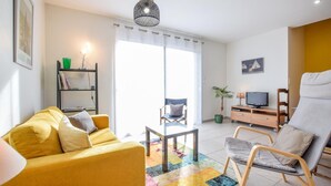 Ein Wohnzimmer mit einem gelben Sofa, einem Holzstuhl, einem Fernseher auf einer Holzkommode und einem farbenfrohen Teppich.