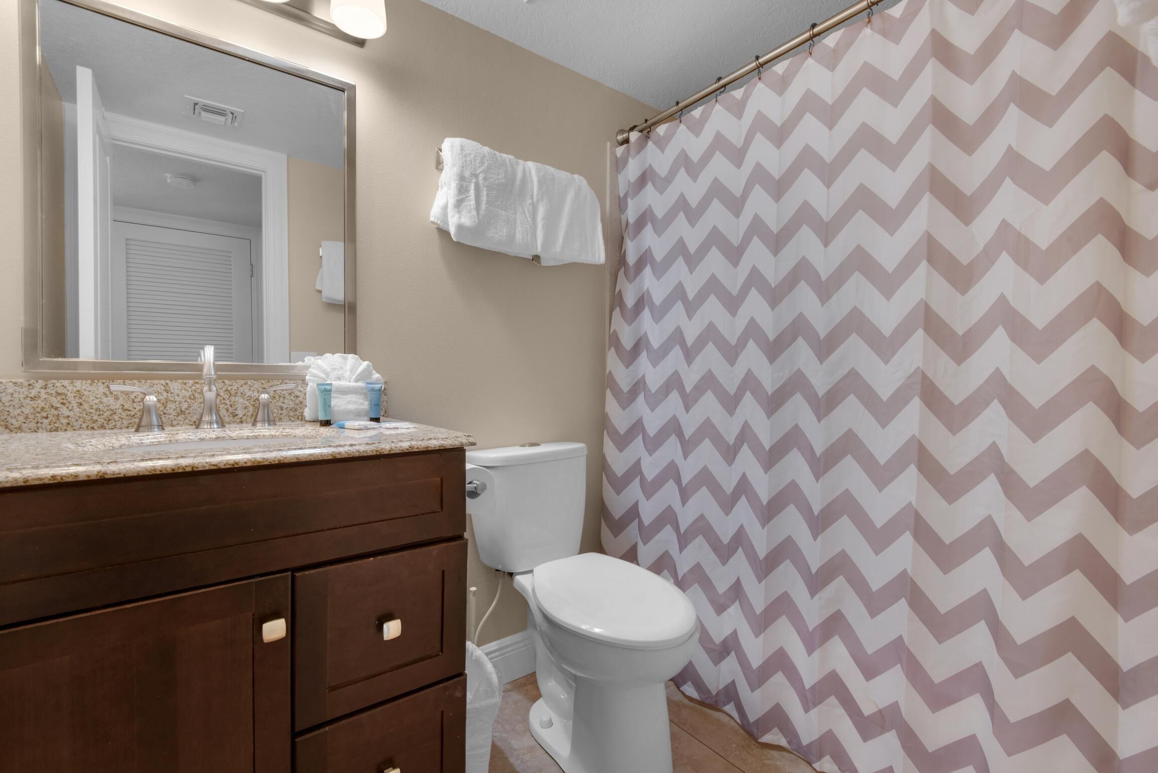 A bathroom with a beige wall, a white toilet, a brown vanity with drawers, a mirror, and a shower curtain with a zigzag pattern.