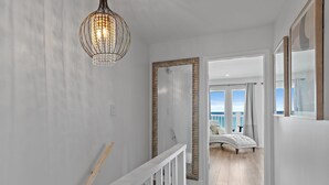 Un couloir avec une rambarde blanche, un luminaire suspendu et une porte donnant sur un balcon où se trouve un canapé blanc.