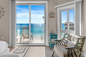 A room with a view of the ocean through a sliding glass door, a white bed, a striped armchair, and a small teal side table. - Empress 1101- Sea Forever (Miramar Beach)