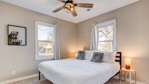 Ein Schlafzimmer mit einem Bett, einem Deckenventilator, zwei Fenstern mit Vorhängen, einem kleinen Nachttisch mit einer Lampe und einem motivierenden Poster an der Wand.