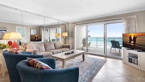 Ein geräumiges Wohnzimmer mit einer Couch, Sesseln, einem Couchtisch und einem Fernseher. Ein Balkon mit Blick auf das Meer ist zu sehen.