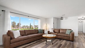 Un salon spacieux, doté d’un grand canapé d’angle, d’une table basse ronde et offrant une vue sur un parc grâce à de grandes fenêtres.