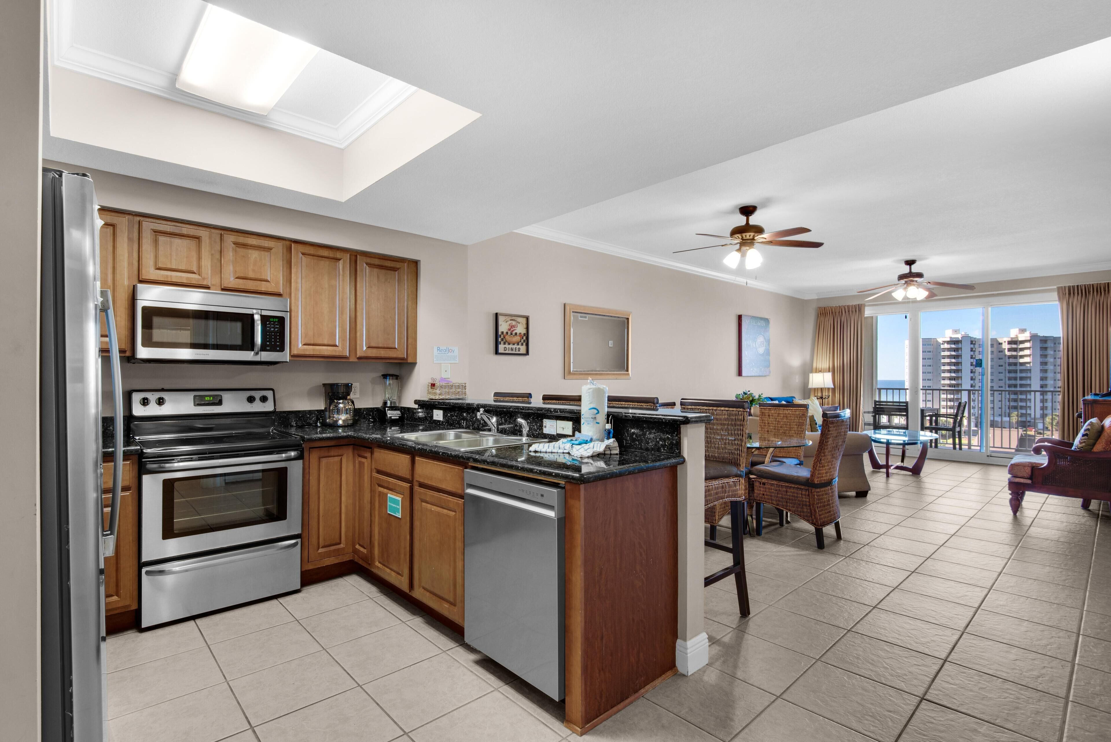 A modern kitchen with stainless steel appliances, wooden cabinets, and a large island. There is a dining area with wicker furniture and a view of the city.