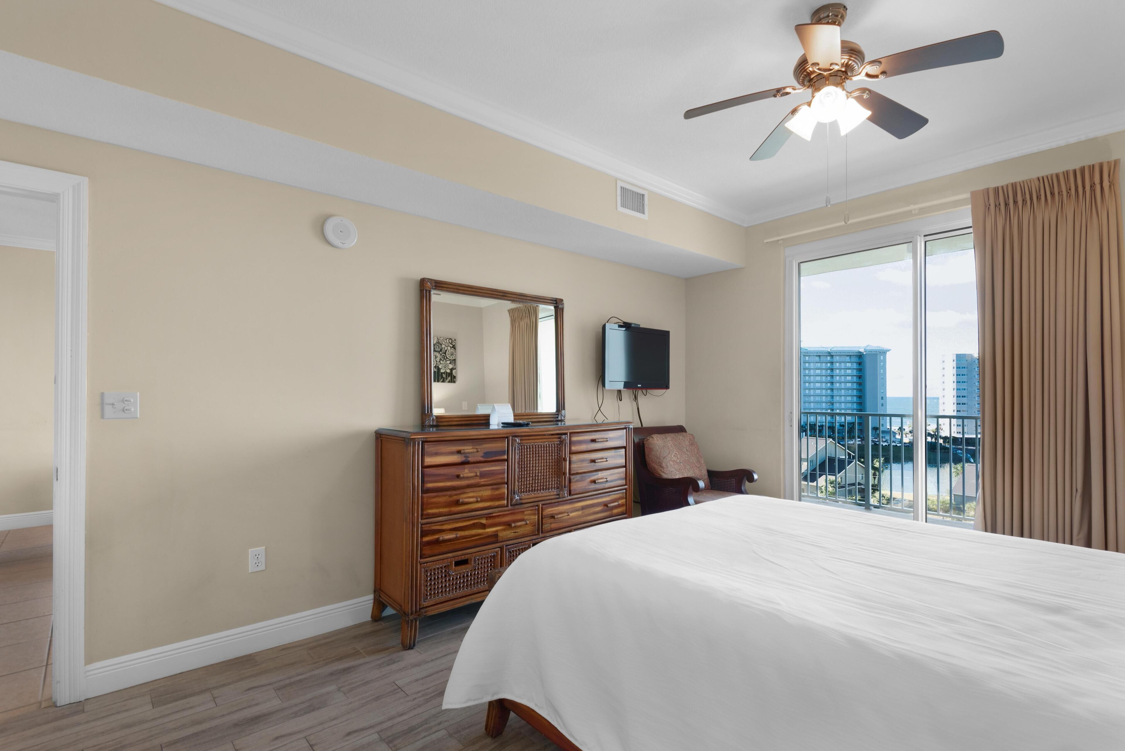 Une chambre d’hôtel dotée d’un grand lit, d’une commode en bois, d’un ventilateur de plafond, d’un téléviseur à écran plat et d’un balcon offrant une vue sur la ville.