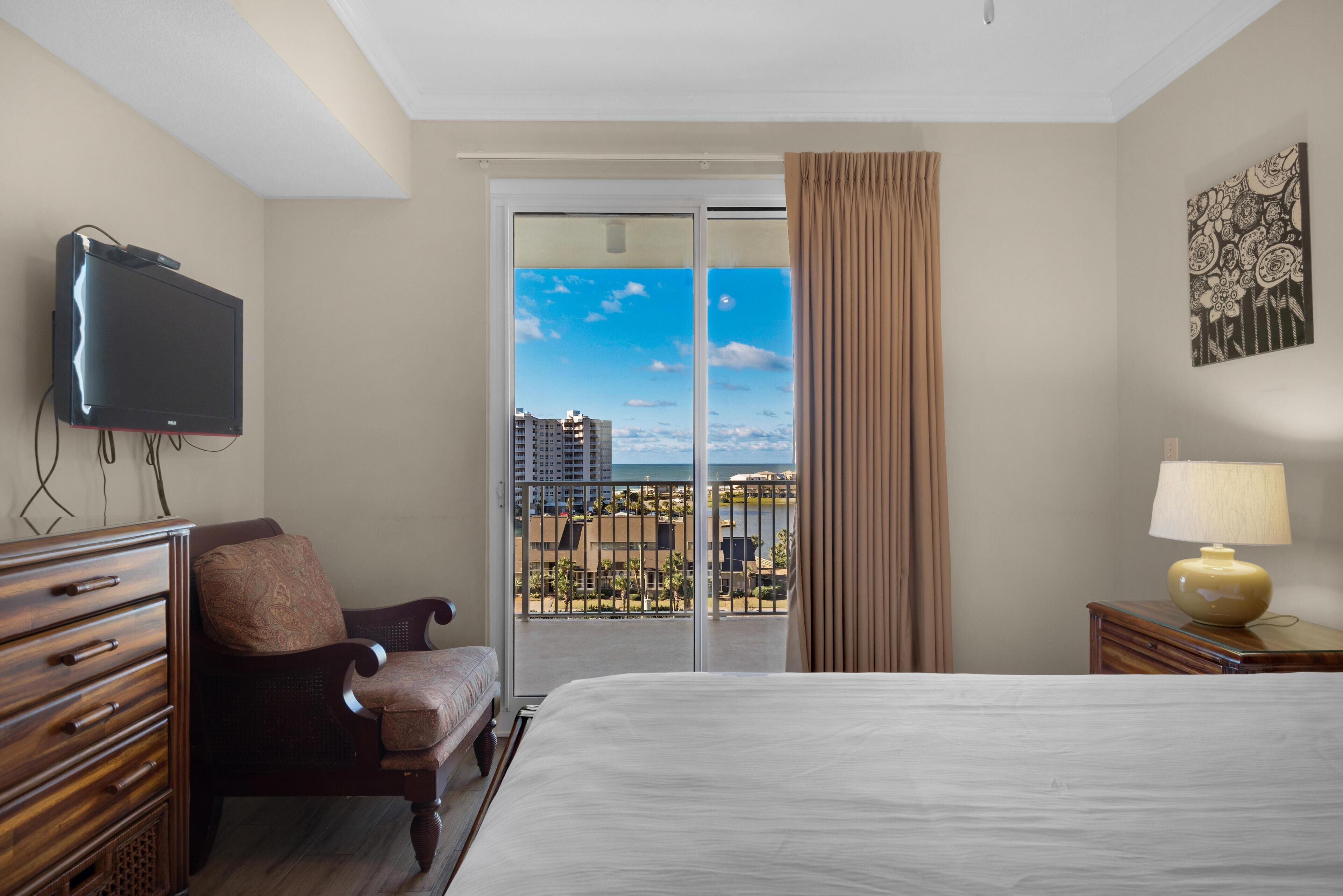 A hotel room with a bed, a chair, a TV, a lamp, and a view of a cityscape through a balcony.