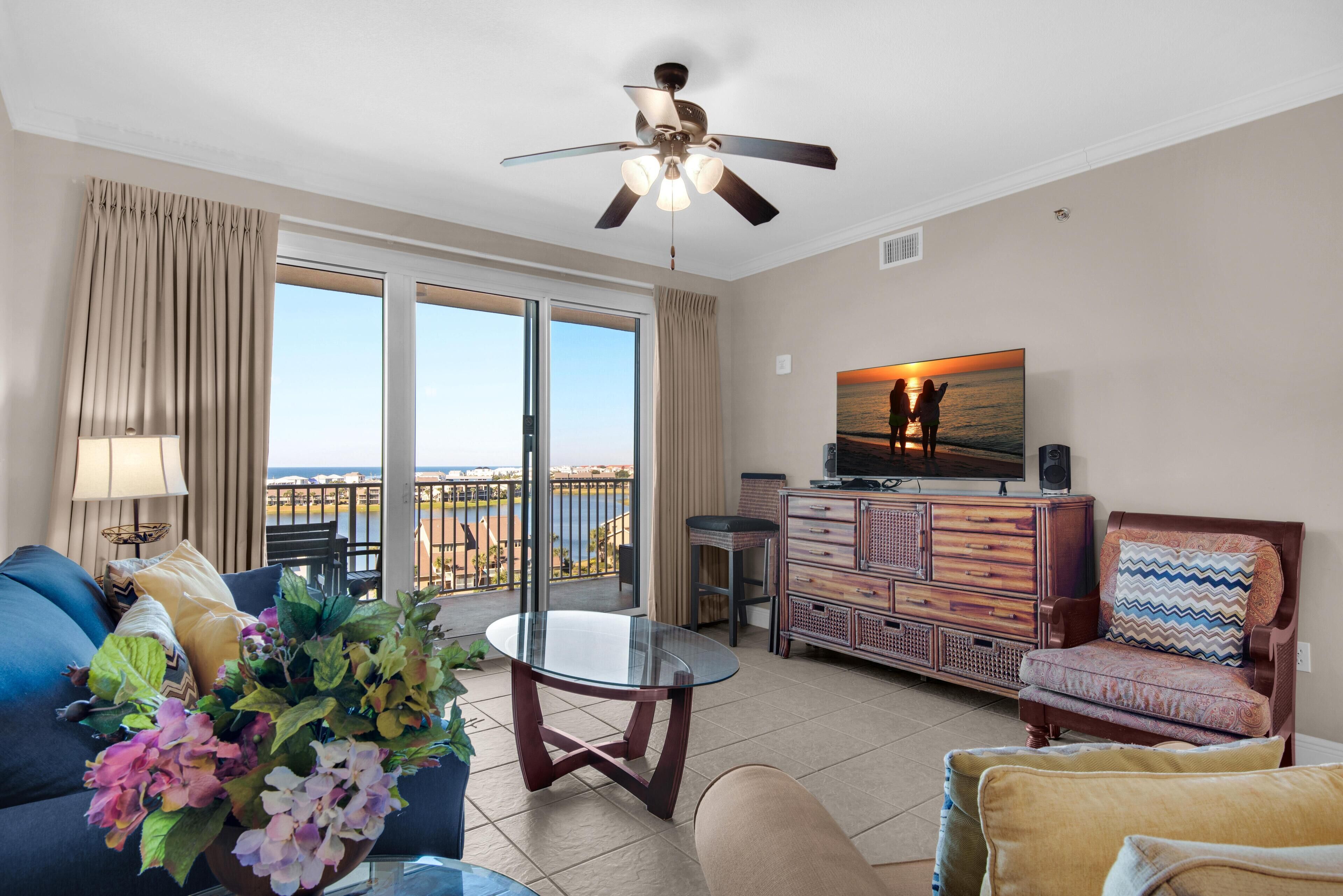 A spacious living room with a balcony, a ceiling fan, a television, and a view of the sea.