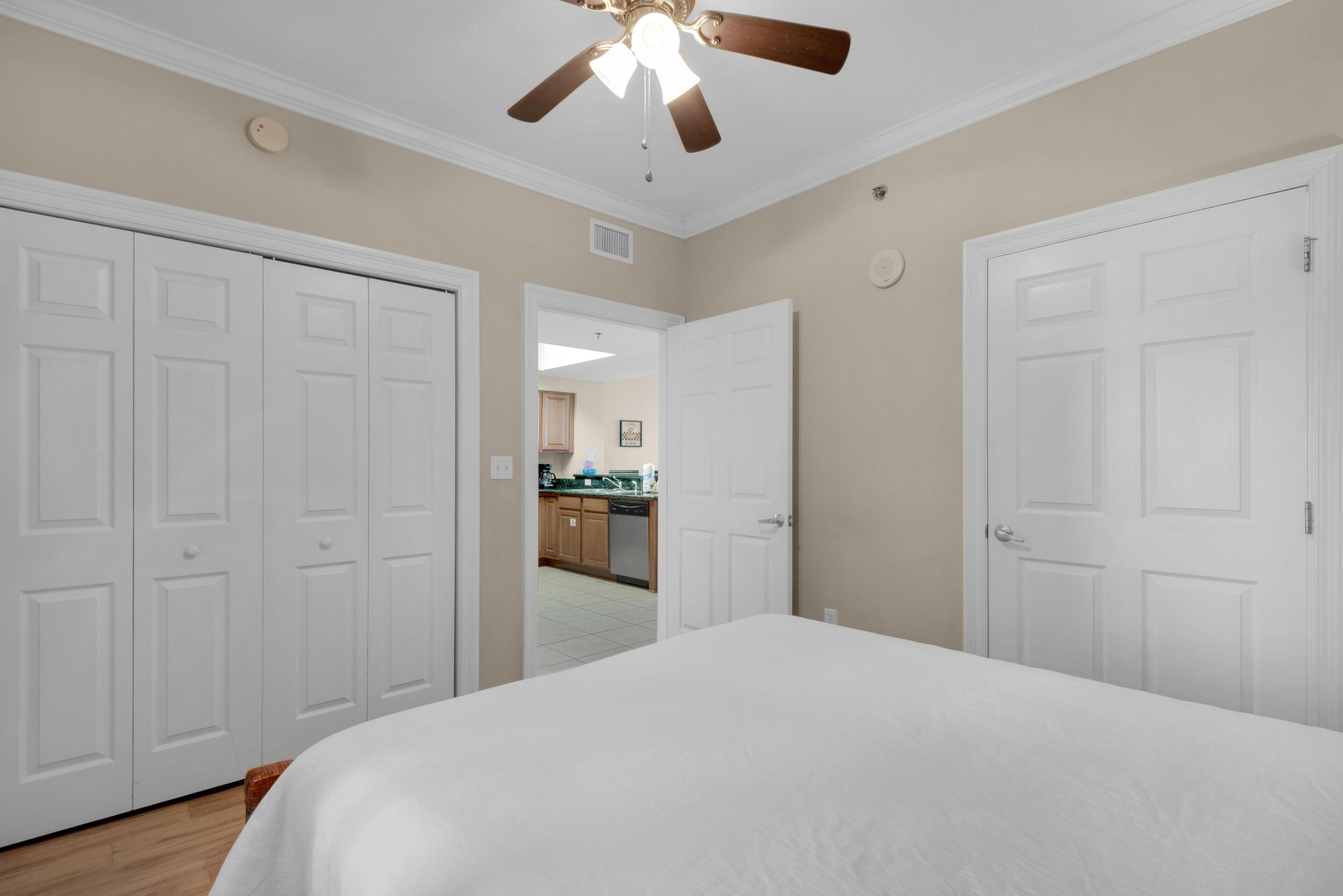 A bedroom with a large bed, white doors, a ceiling fan, and a view into the kitchen.