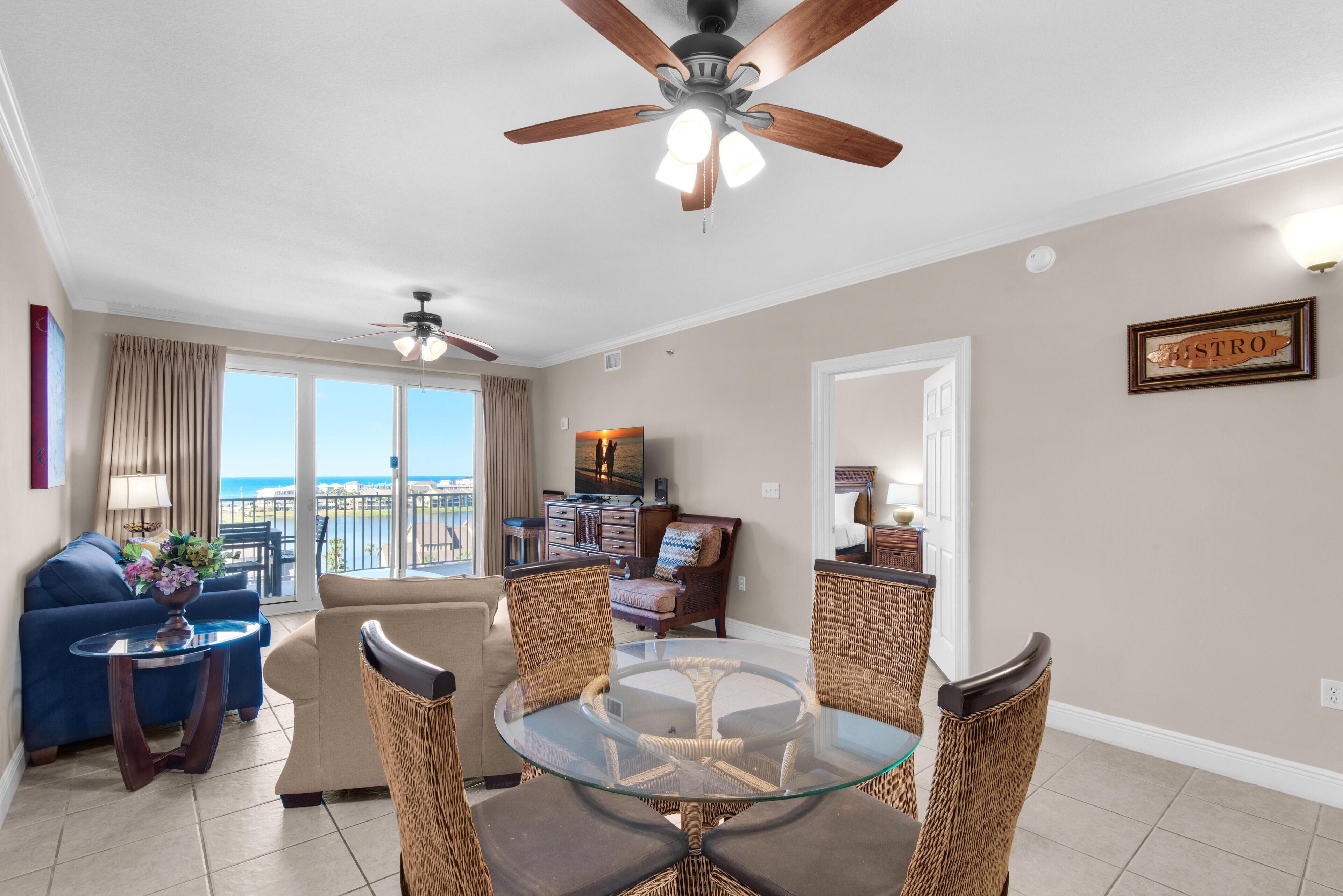 A spacious living area with a glass-top dining table surrounded by wicker chairs, a ceiling fan, and a view of the ocean through large windows.